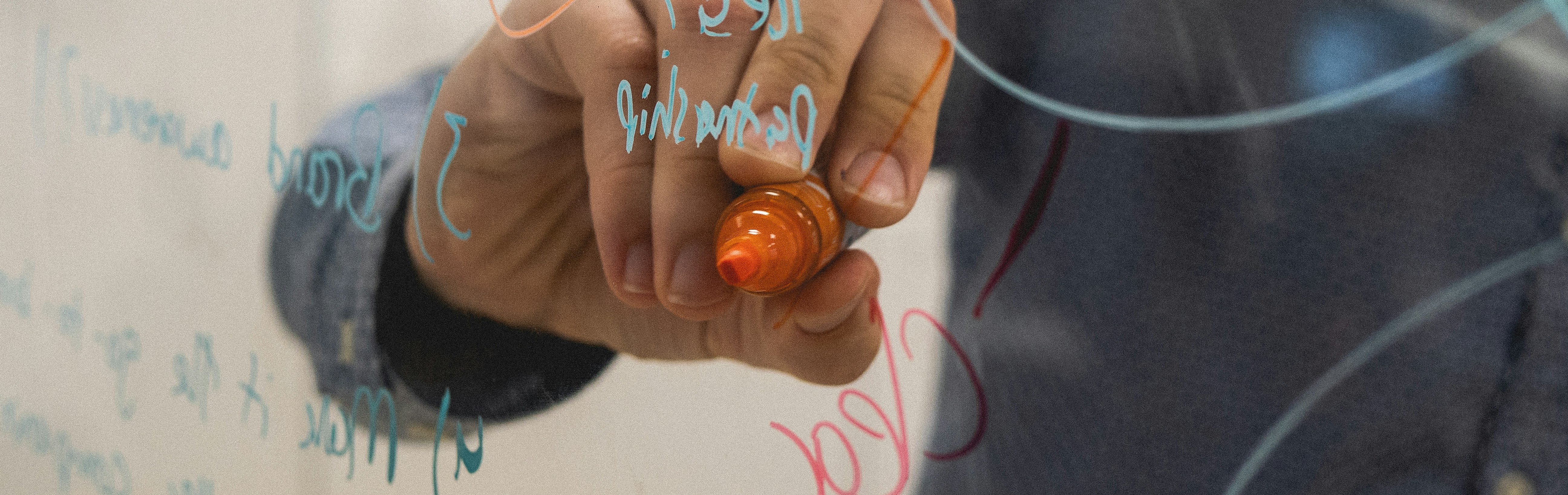 Hand schreibt mit orangenem Stift auf einer durchsichtigen Plexiglasscheibe