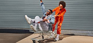 Eine junger Mann schiebt einen anderen jungen Mann im Einkaufswagen