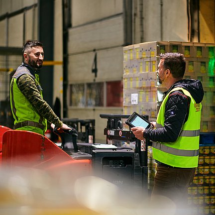 Zwei Männer mit Sicherheitswesten im Lager beladen einen LKW mit Pallette